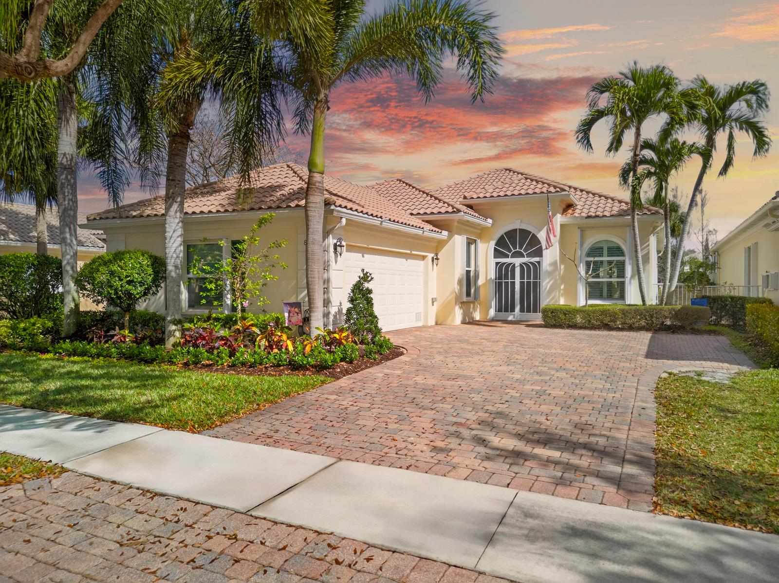 A single-story house with a tiled roof, brick paver driveway, and palm trees at sunset — 8223 Pine Cay Road, Wellington FL 33414, WELLINGTON, FL
