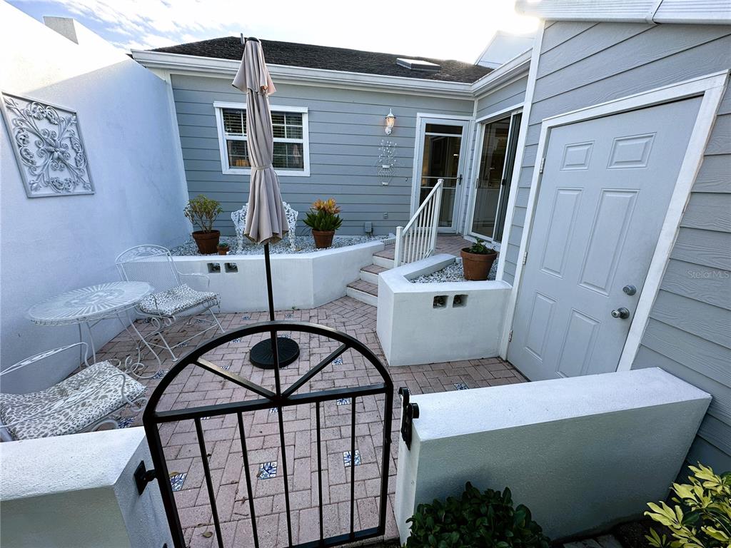 A paved courtyard features a metal gate, patio furniture, a closed umbrella, and white walls — 517 WOODSTORK CIRCLE, BRADENTON, FL