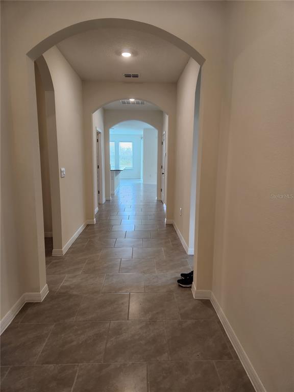 A long hallway with arched entryways and light-colored tile flooring leads toward a bright room — 6930 AUDOBON OSPREY COVE, HARMONY, FL