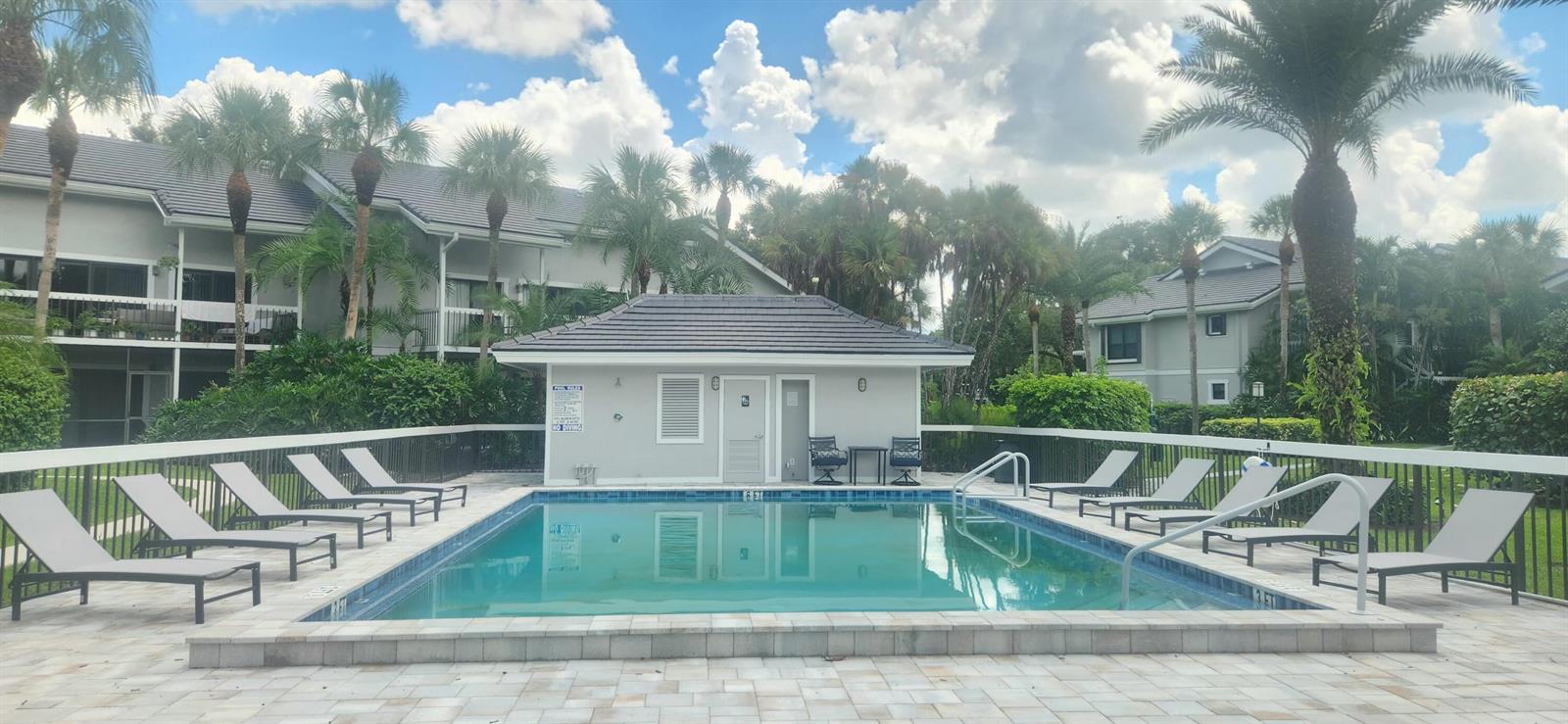 A rectangular swimming pool is surrounded by lounge chairs and a small building with palm trees — 11863 Wimbledon Circle # 416, Wellington FL 33414, WELLINGTON, FL