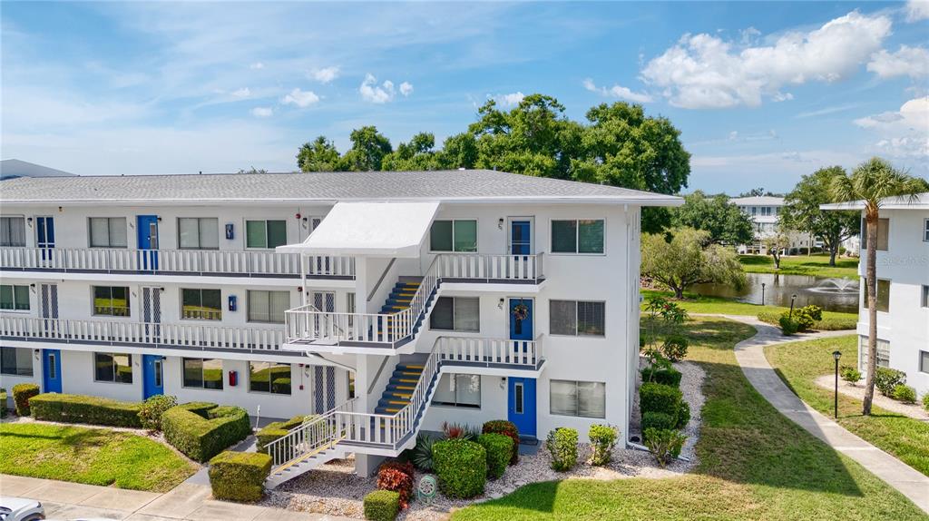 A three-story white apartment building features exterior staircases, blue doors, and a nearby pond — 11251 80TH AVENUE UNIT 202, SEMINOLE, FL