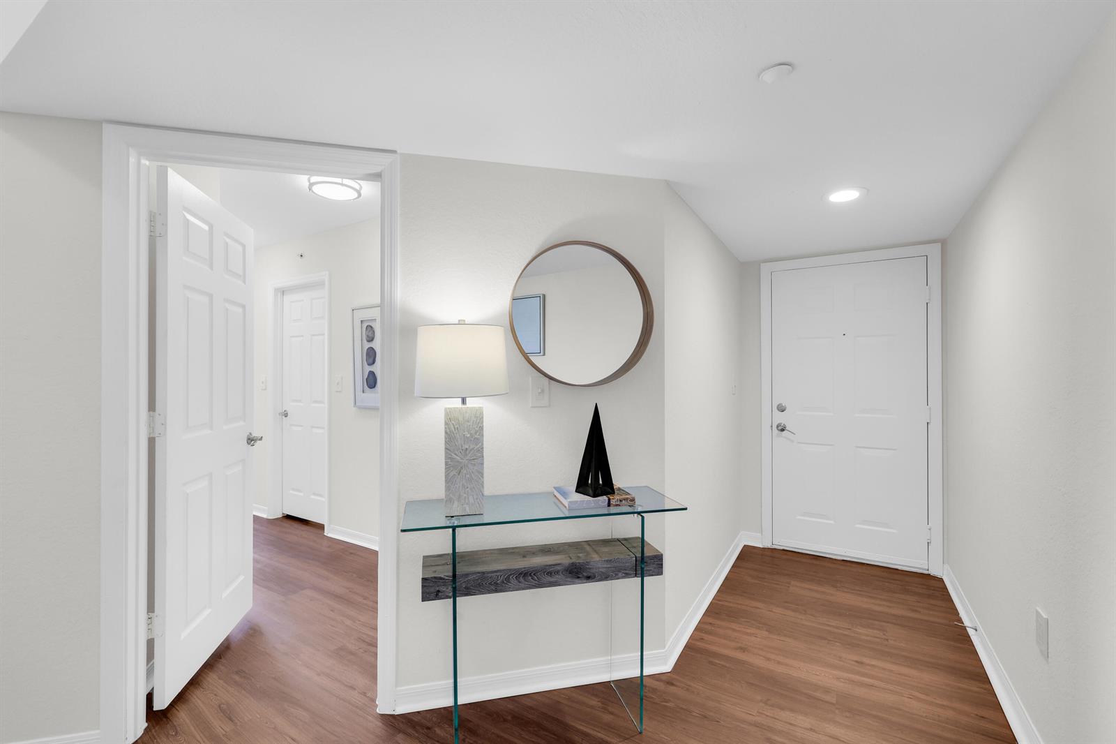 A bright entryway features wood-look flooring, a glass console table, a round mirror, and doors — 15035 Michelangelo Boulevard # 303, Delray Beach FL 33446, DELRAY BEACH, FL