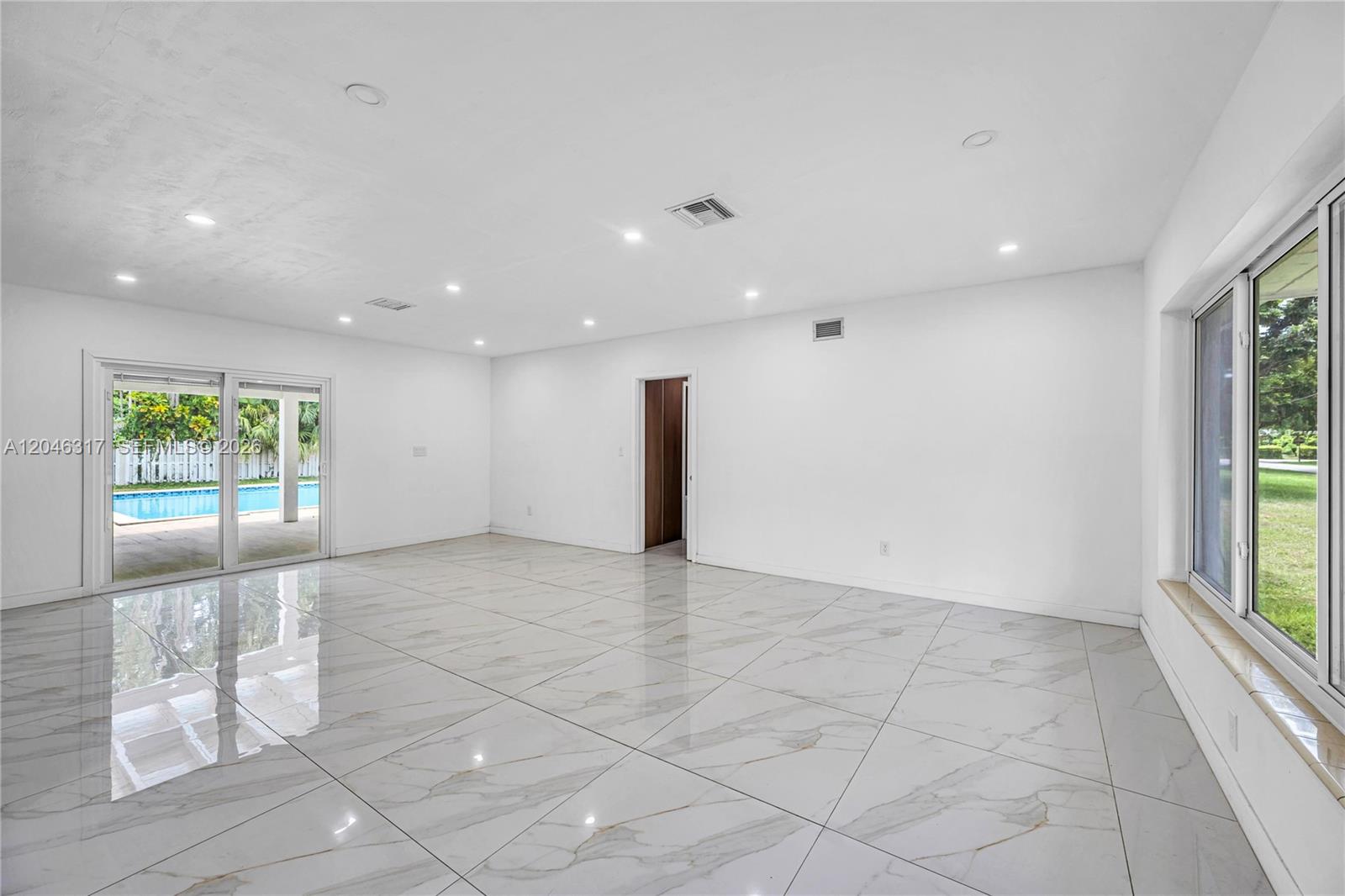 A bright, empty living room features white marble-look tile floors and sliding glass patio doors — 12525 SW 68th Ct, Pinecrest FL 33156, PINECREST, FL
