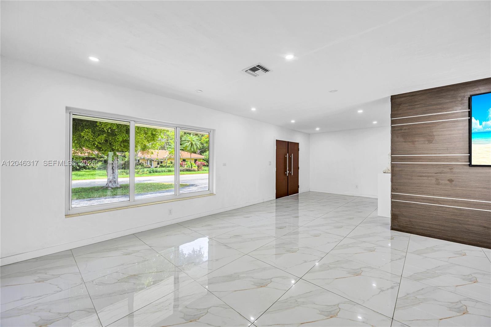 A bright, empty room features white marble floors, a large window, and a wood-paneled wall — 12525 SW 68th Ct, Pinecrest FL 33156, PINECREST, FL