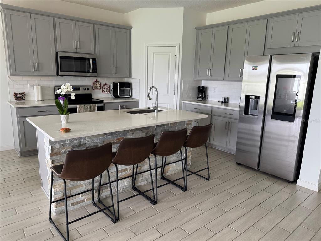 A kitchen features gray cabinets, a stone-front island with four stools, and stainless steel appliances — 1177 TUPELO TRAIL, HAINES CITY, FL