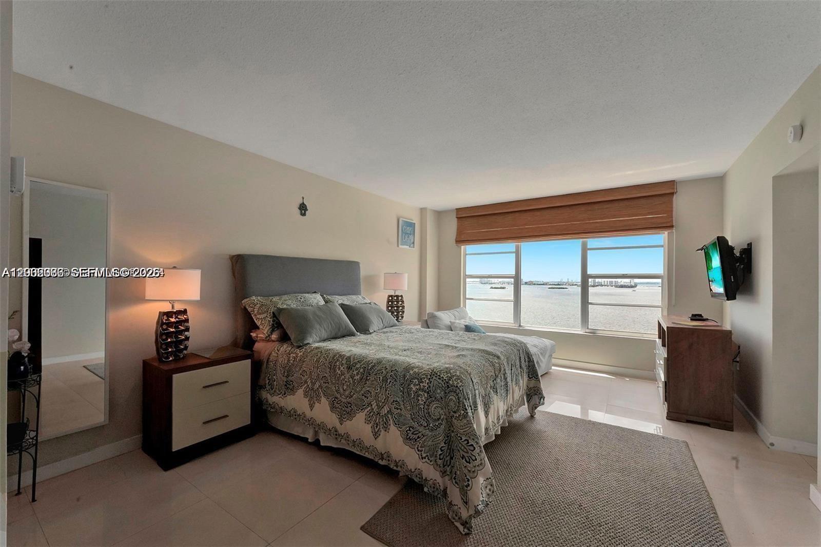 A bedroom features a bed, nightstand, television, and a large window overlooking the ocean — 11111 Biscayne Blvd # 18E, Miami FL 33181, MIAMI, FL