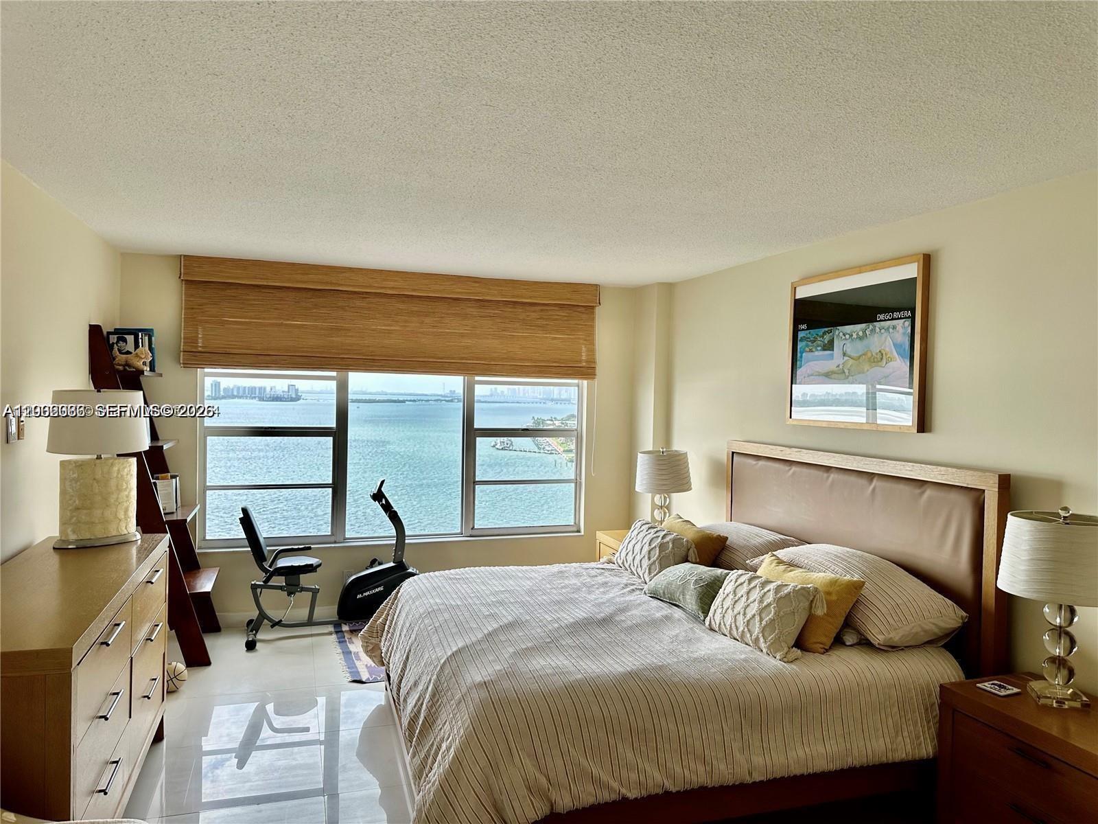 A bedroom features a king bed, a dresser, an exercise bike, and a waterfront view — 11111 Biscayne Blvd # 18E, Miami FL 33181, MIAMI, FL