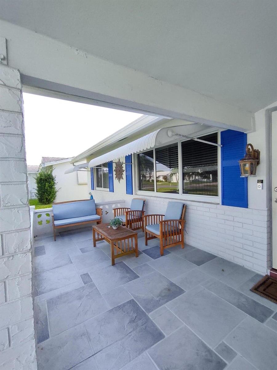 A covered patio features gray stone-look flooring, white brick walls, and blue-cushioned wooden outdoor furniture — 1826 SW Congress Boulevard, Boynton Beach FL 33426, BOYNTON BEACH, FL