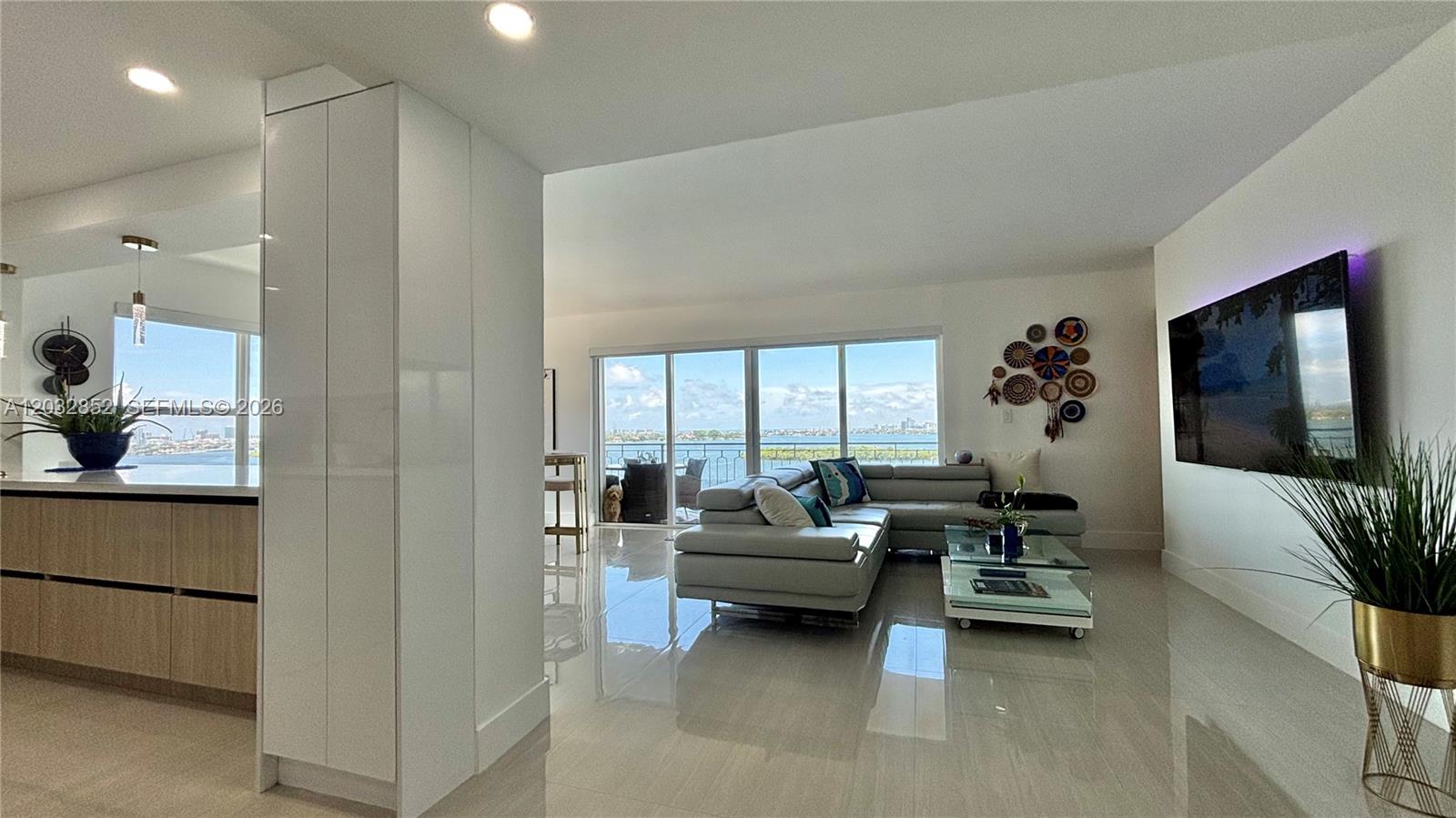 A modern living room features a grey sectional sofa, glass coffee table, and waterfront views — 11111 NE Biscayne Blvd # 16A, Miami FL 33181, MIAMI, FL