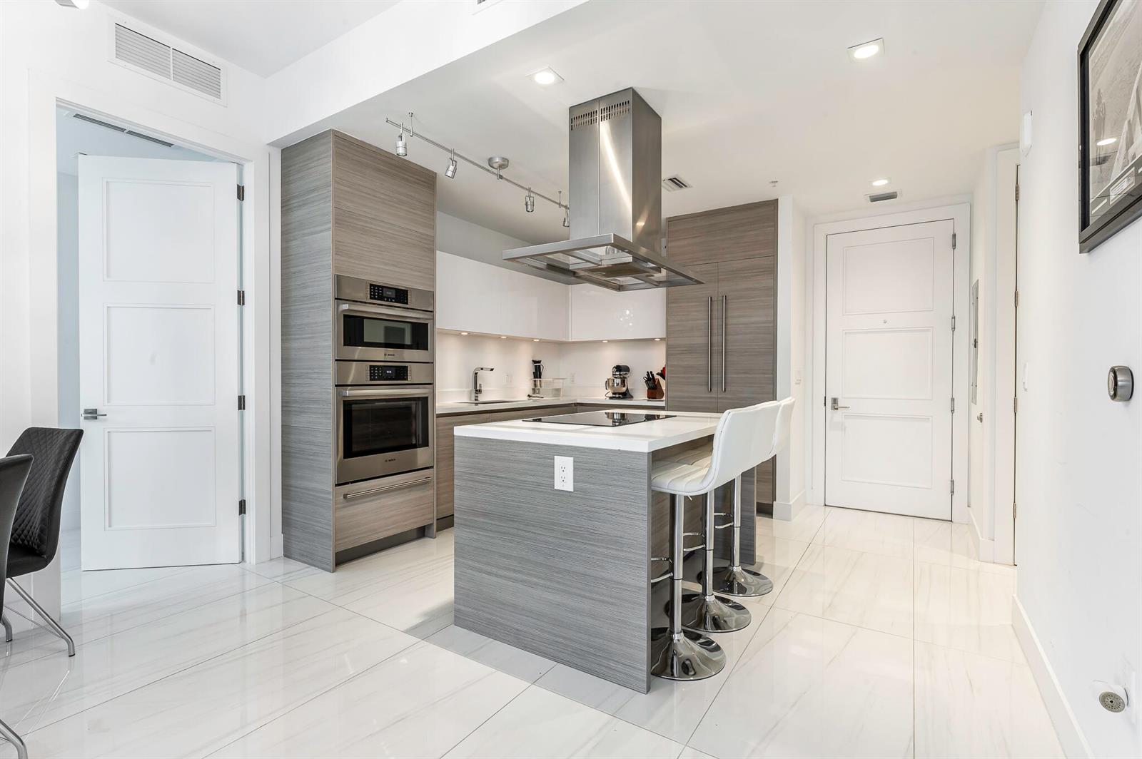 A modern kitchen features gray wood-grain cabinetry, white countertops, a central island, and stainless appliances — 155 E Boca Raton Road # 415, Boca Raton FL 33432, BOCA RATON, FL