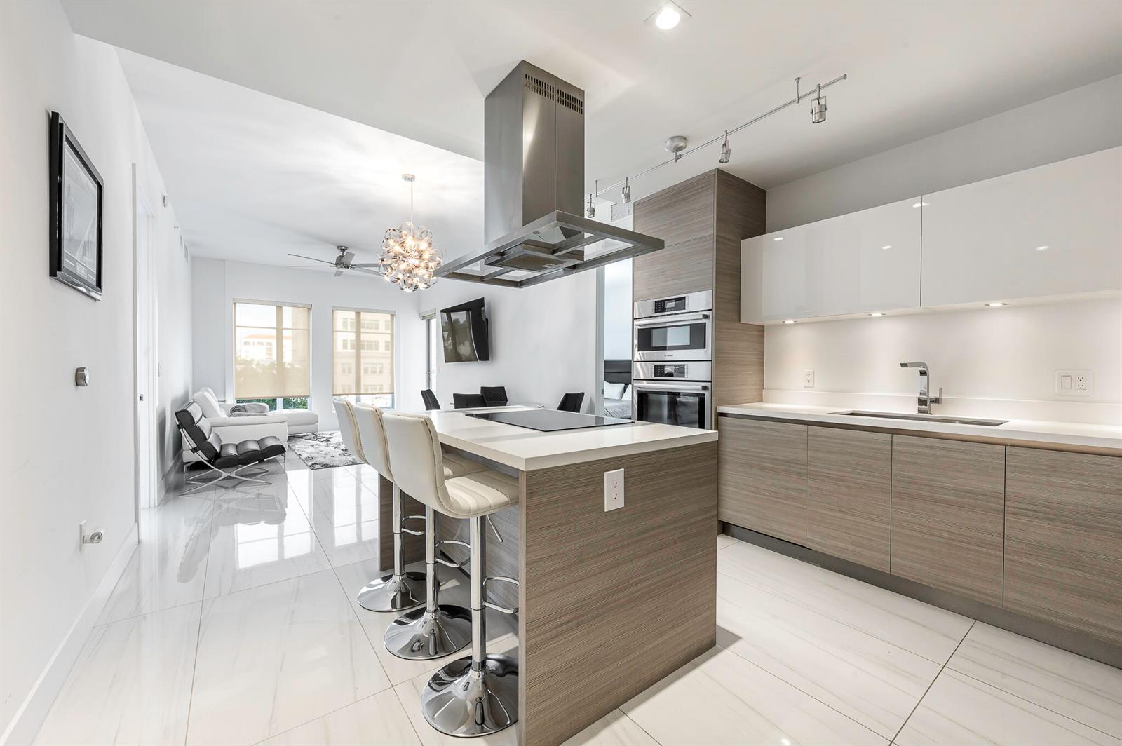 A modern kitchen features wood-grain cabinetry, a central island with bar stools, and white flooring — 155 E Boca Raton Road # 415, Boca Raton FL 33432, BOCA RATON, FL