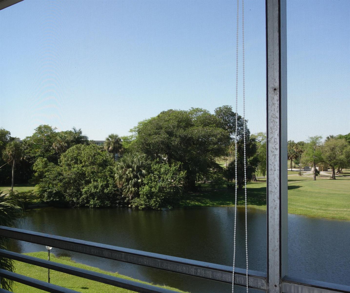 A view of a pond and trees seen through a screened-in balcony railing — 3507 Oaks Way # 307, Pompano Beach FL 33069, POMPANO BEACH, FL