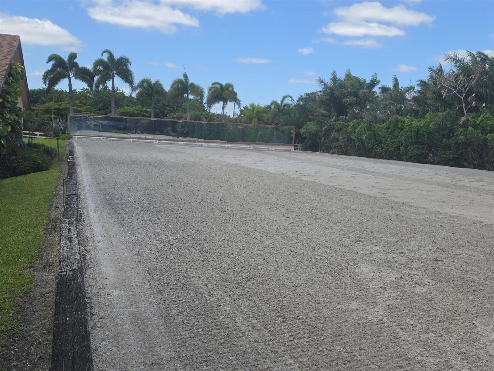 A large, outdoor sand riding arena is bordered by a wooden fence and palm trees — 14127 Equestrian Way, Wellington FL 33414, WELLINGTON, FL