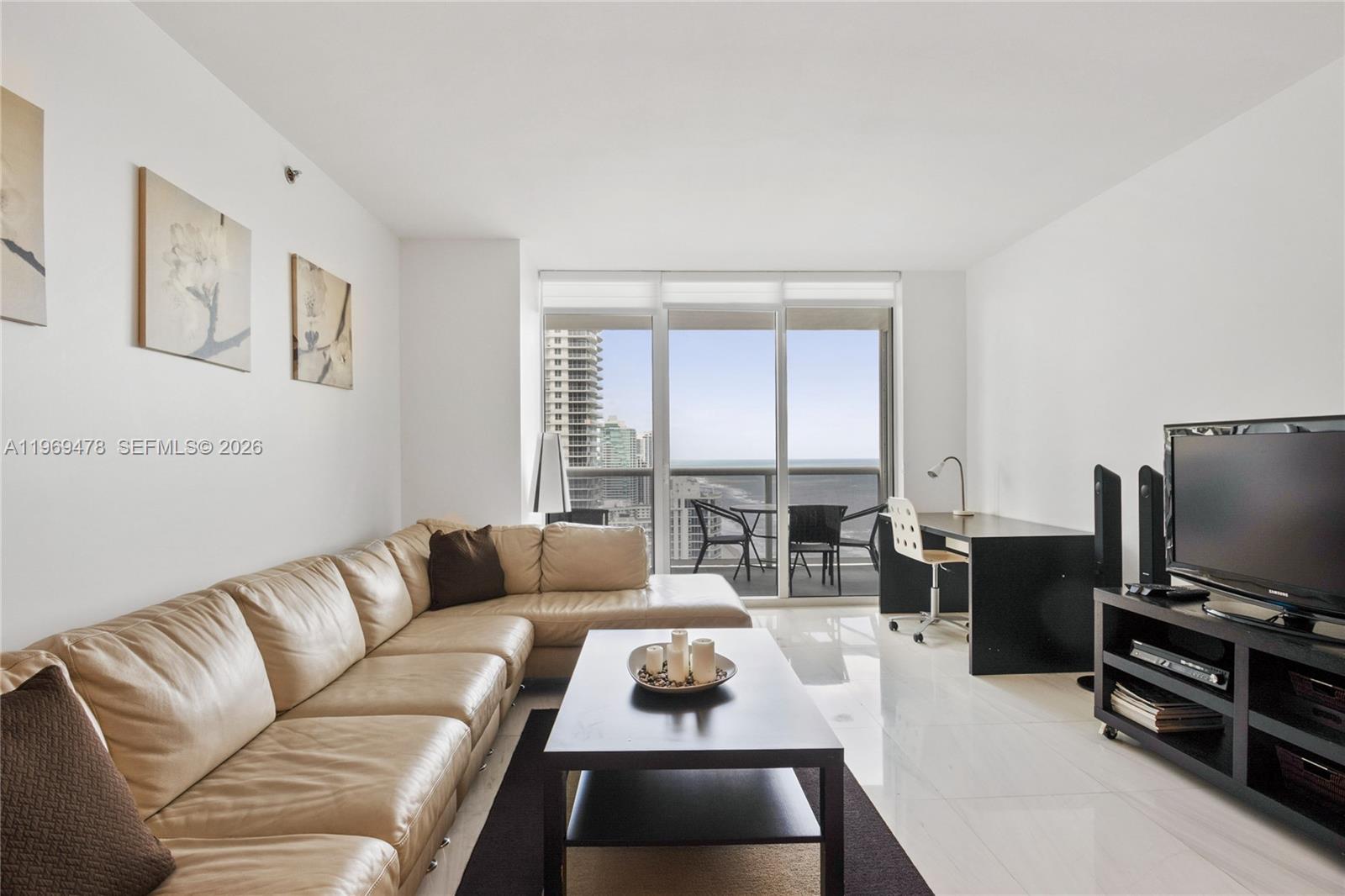 A living room features a beige sectional sofa, dark coffee table, desk, and balcony ocean views — 1830 S OCEAN DR # 3211, Hallandale Beach FL 33009, HALLANDALE BEACH, FL