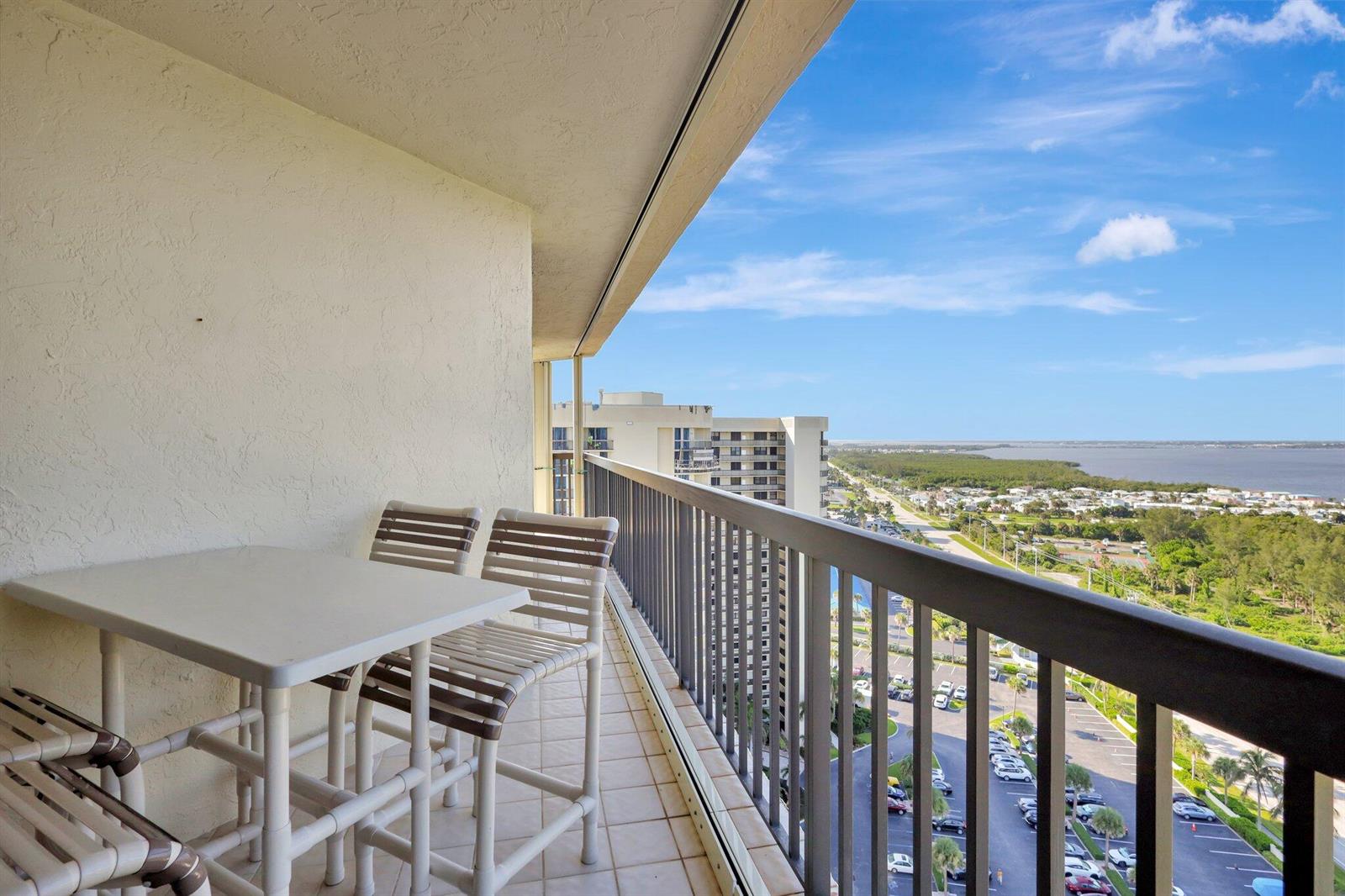 A high-rise balcony with patio furniture overlooks a parking lot, trees, and a distant bay — 9500 S Ocean Drive # 10, Jensen Beach FL 34957, JENSEN BEACH, FL