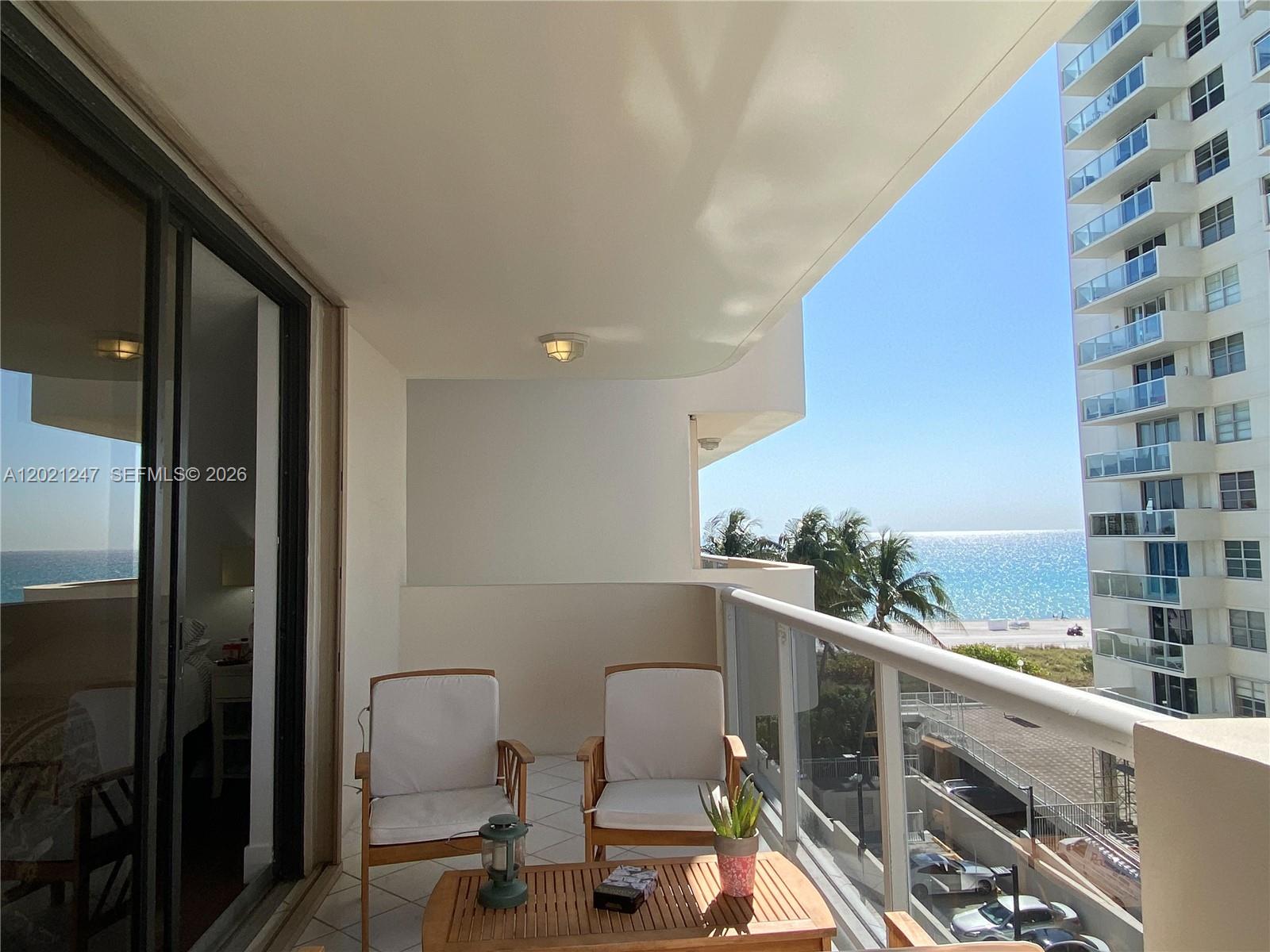 A balcony with two chairs and a table overlooks the ocean and a neighboring building — 5757 Collins Ave # 703, Miami Beach FL 33140, MIAMI BEACH, FL