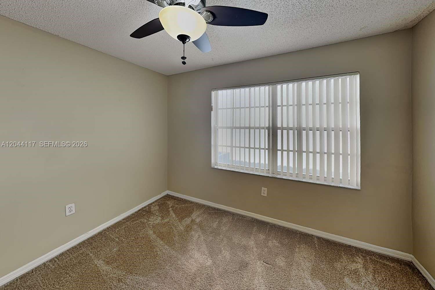 An empty bedroom features beige walls, tan carpeting, a ceiling fan, and vertical window blinds — PLANTATION, FL