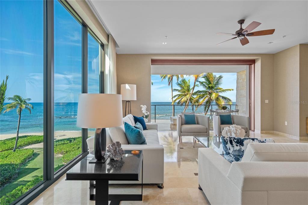 A bright living room features floor-to-ceiling windows and a balcony overlooking the ocean and palms — 200 Dorado Beach Drive 3731 WEST BEACH, A RITZ-CARLTON RESIDENCES UNIT 3731, DORADO, PR