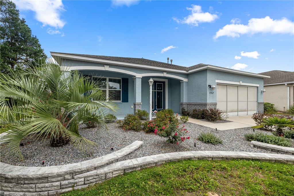 A light blue single-story house features a covered entryway, a garage, and landscaped gravel beds — 7767 SW 80TH PLACE ROAD, OCALA, FL