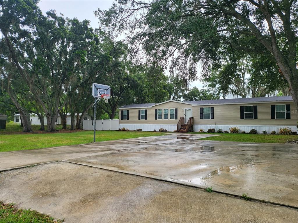A single-story manufactured home with tan siding sits behind a large concrete driveway and basketball hoop — 8915 HAMMOCK LOOP, POLK CITY, FL 33868, POLK CITY, FL