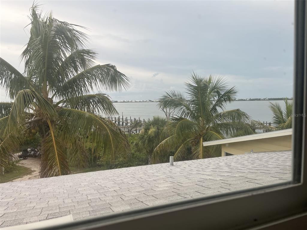 A view from a window overlooking a shingled roof, palm trees, and a distant dock — 9400 LITTLE GASPARILLA ISLAND UNIT A7, PLACIDA, FL