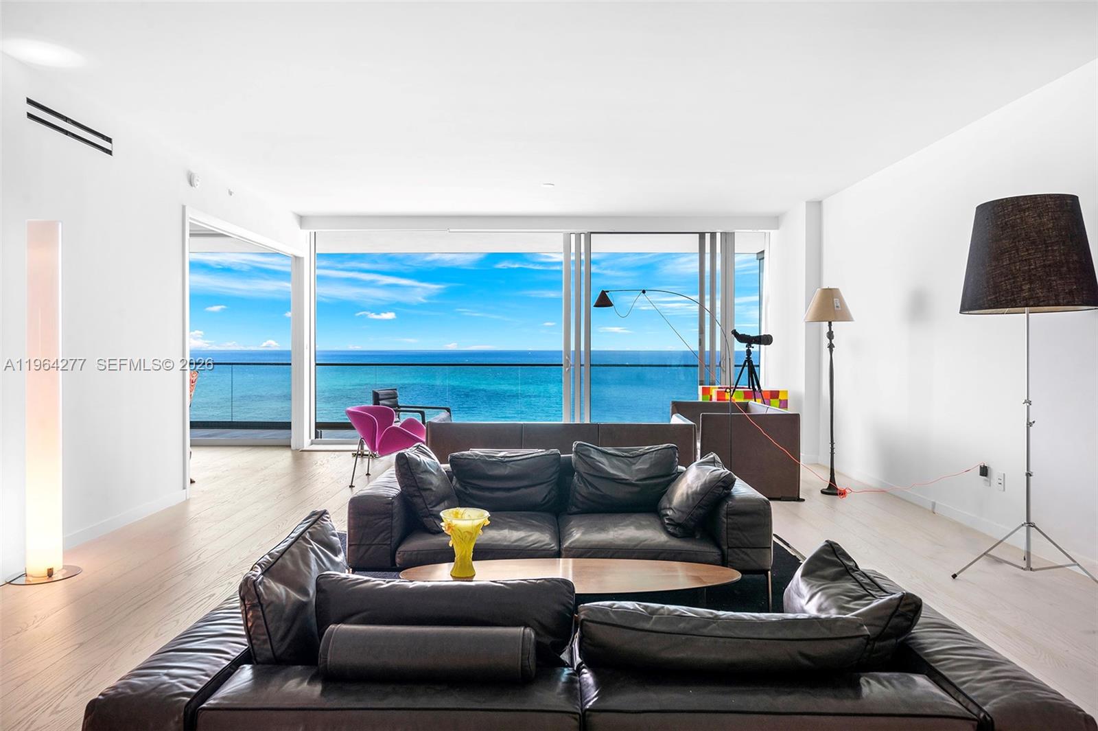 A modern living room features dark leather sofas, light wood floors, and floor-to-ceiling ocean views — 10203 Collins Ave # 1602, Bal Harbour FL 33154, BAL HARBOUR, FL