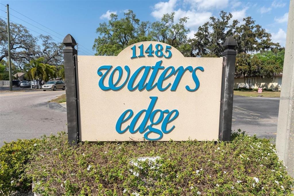 A sign for a community named Waters Edge stands outdoors near a road and landscaping — 11485 OAKHURST ROAD UNIT 200-307, LARGO, FL