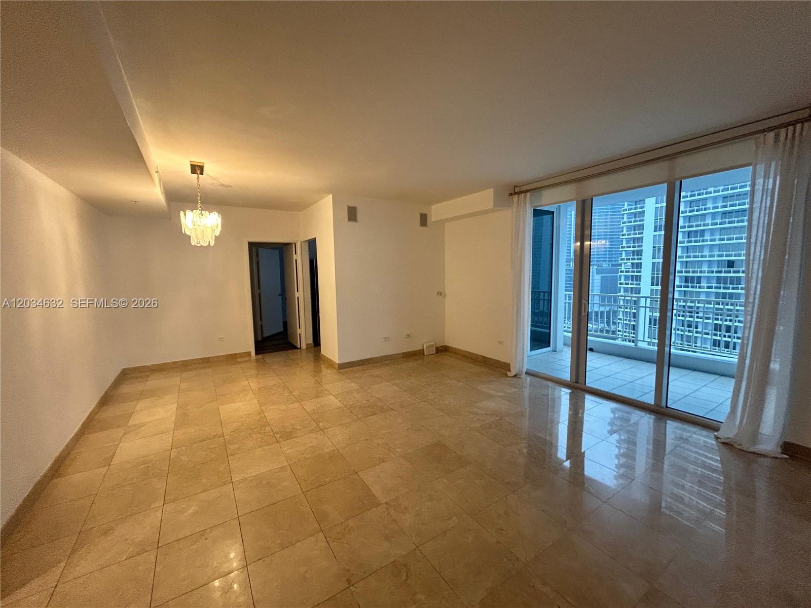 An empty living room features beige tile flooring, a crystal chandelier, and a glass balcony door — 801 Brickell Key Blvd # 1710, Miami FL 33131, MIAMI, FL