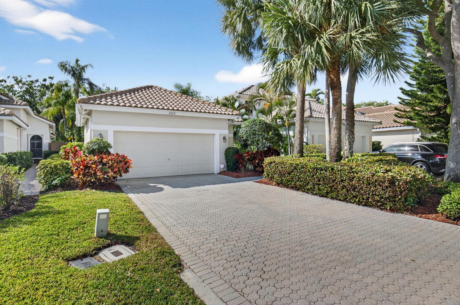 A beige house with a tiled roof and a paved driveway sits behind a manicured hedge — 2476 NW 66th Drive, Boca Raton FL 33496, BOCA RATON, FL