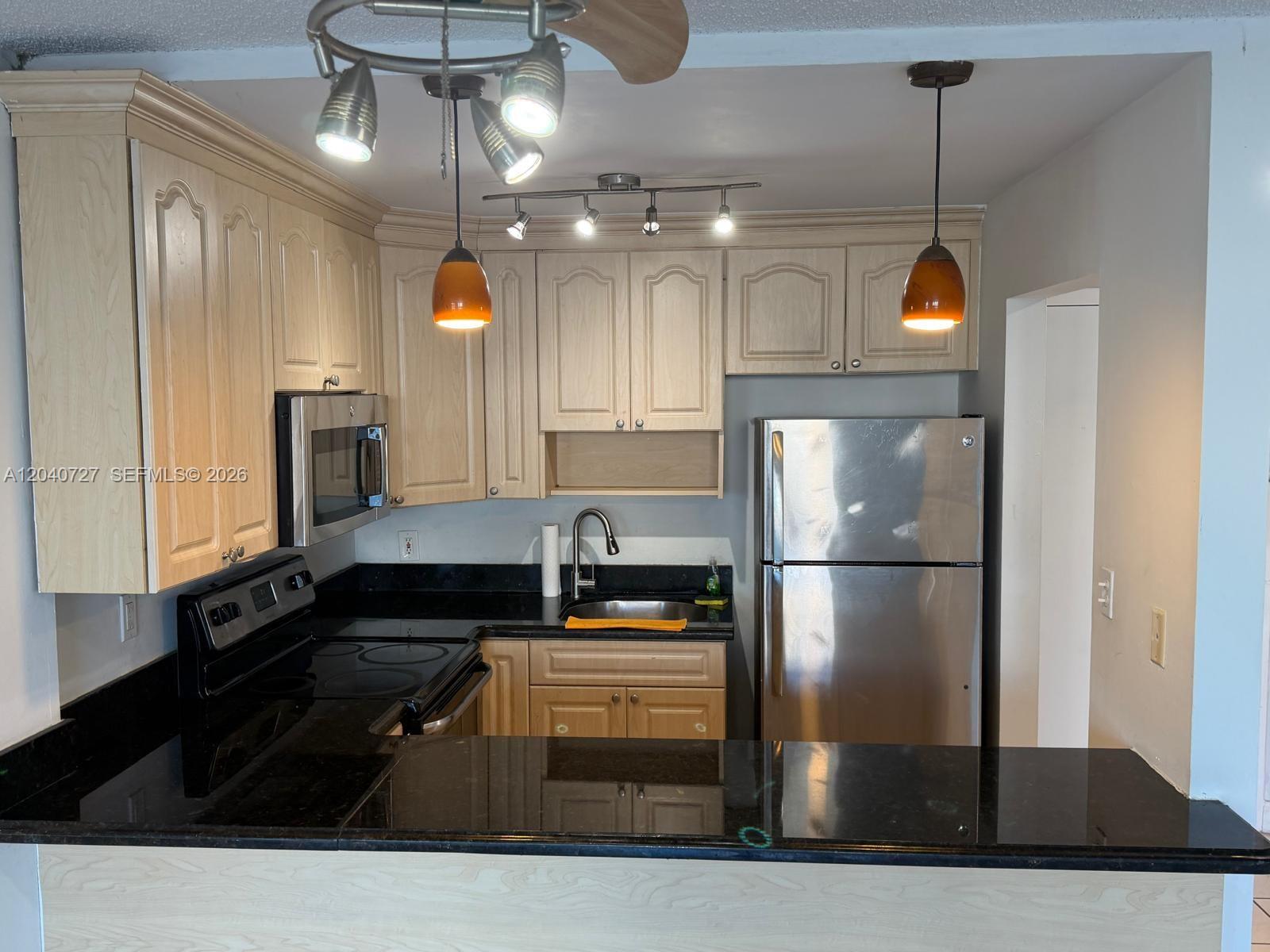 A kitchen features light wood cabinets, black countertops, stainless steel appliances, and pendant lighting — 1990 Marseille Dr # 303, Miami Beach FL 33141, MIAMI BEACH, FL