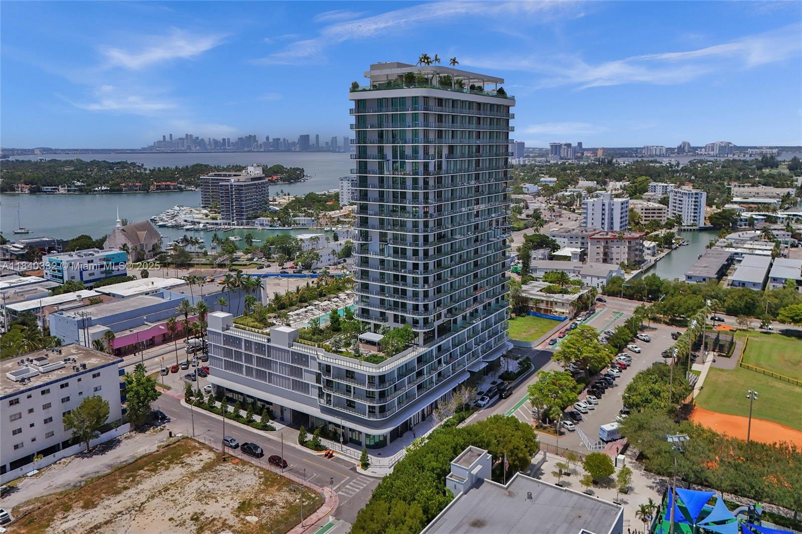 An aerial view shows a modern high-rise residential building surrounded by urban streets and water — 580 72nd St # 1208, Miami Beach FL 33141, MIAMI BEACH, FL