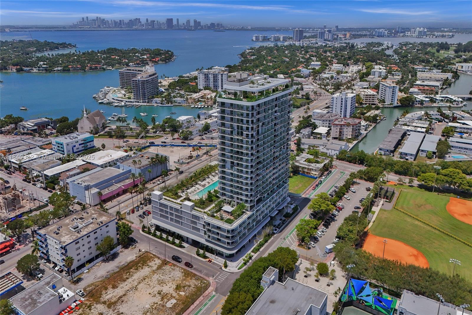 An aerial view shows a modern high-rise residential building surrounded by urban streets and water — 580 72nd St # 1208, Miami Beach FL 33141, MIAMI BEACH, FL