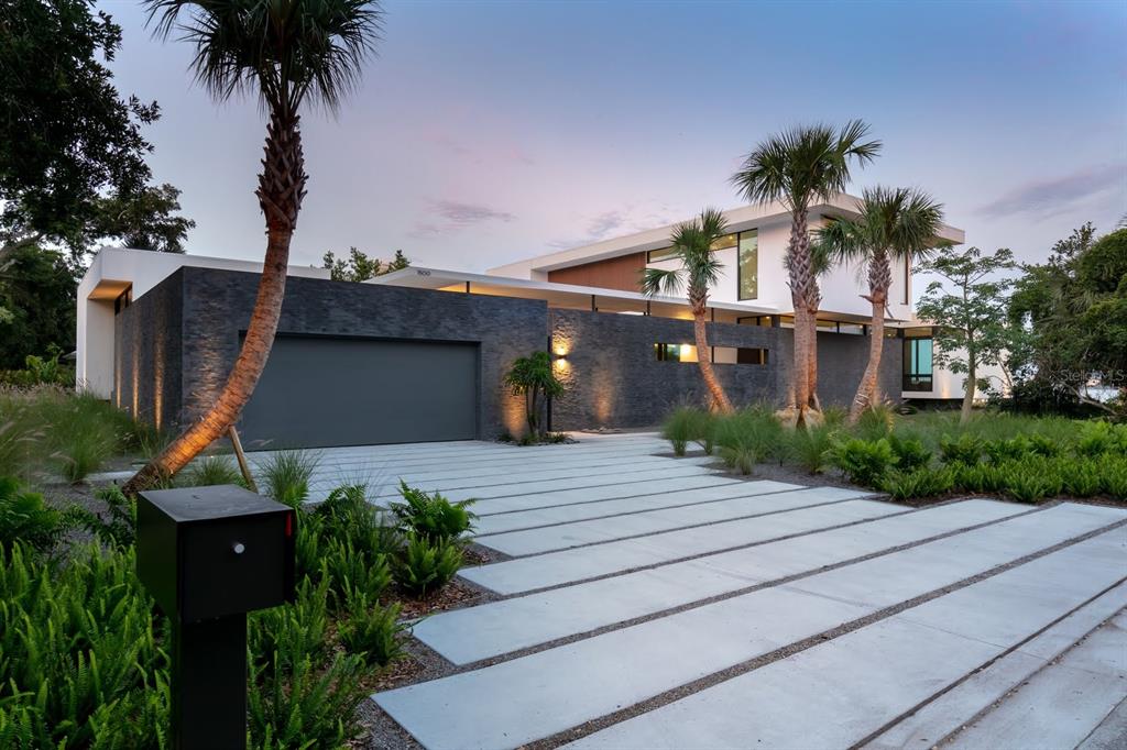 A modern house with a dark stone facade, concrete driveway, and palm trees at dusk — 1500 BAYVIEW DRIVE, SARASOTA, FL