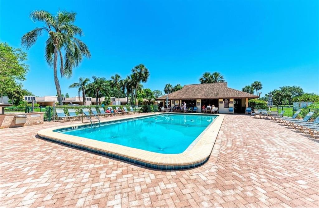 A rectangular swimming pool sits on a brick patio surrounded by palm trees and buildings — 6116 43RD STREET W UNIT 401D, BRADENTON, FL