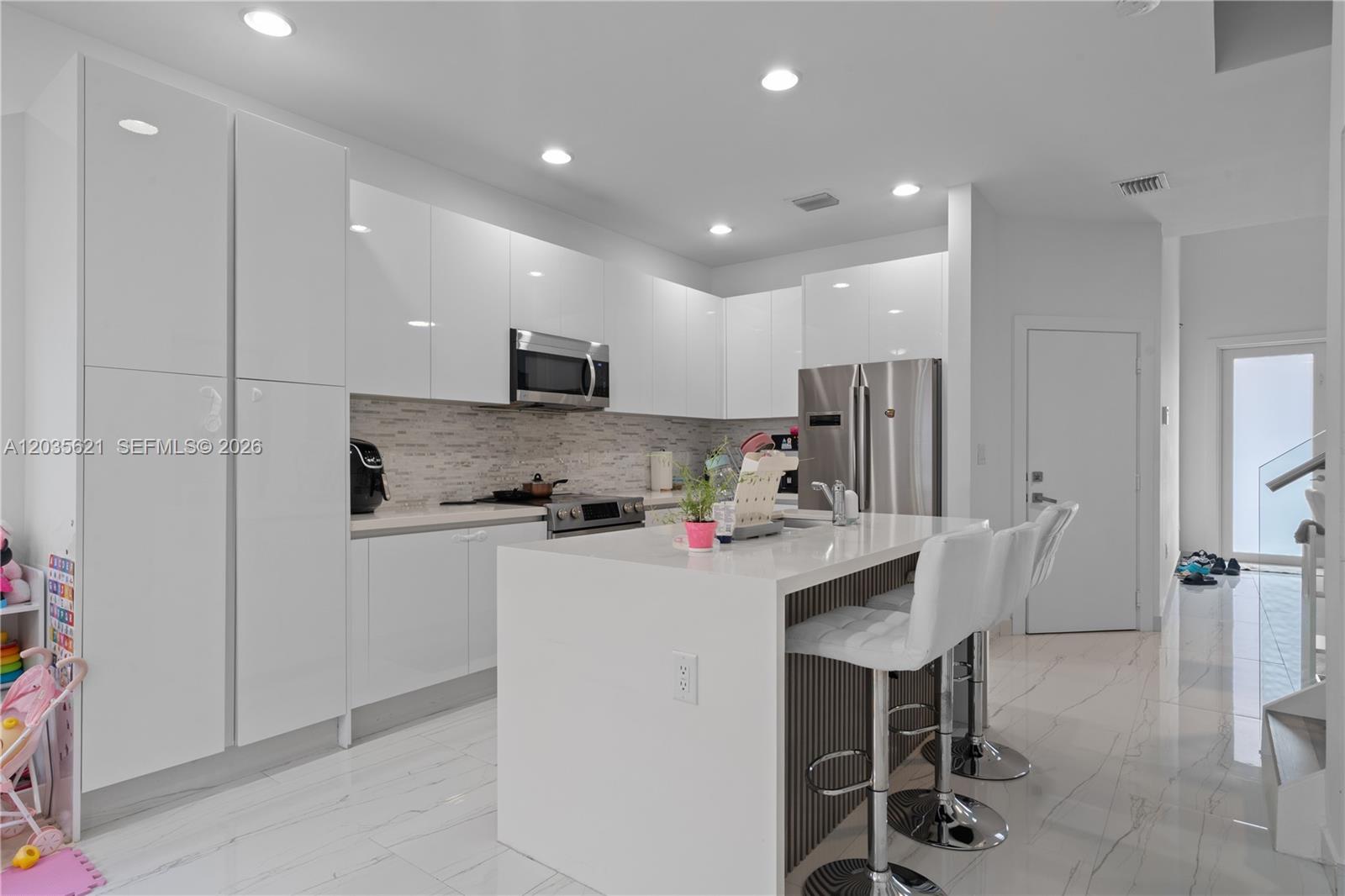 A modern kitchen features white glossy cabinets, a central island with bar seating, and tile flooring — 10294 NW 71st Ter, Doral FL 33178, DORAL, FL