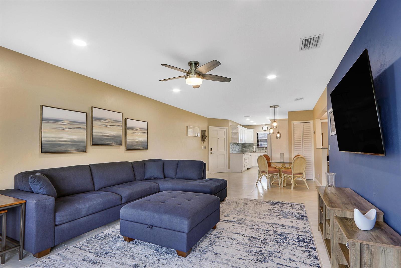 A living room features a blue sectional sofa, a matching ottoman, and a wall-mounted television — 755 Saturn Street # E204, Jupiter FL 33477, JUPITER, FL