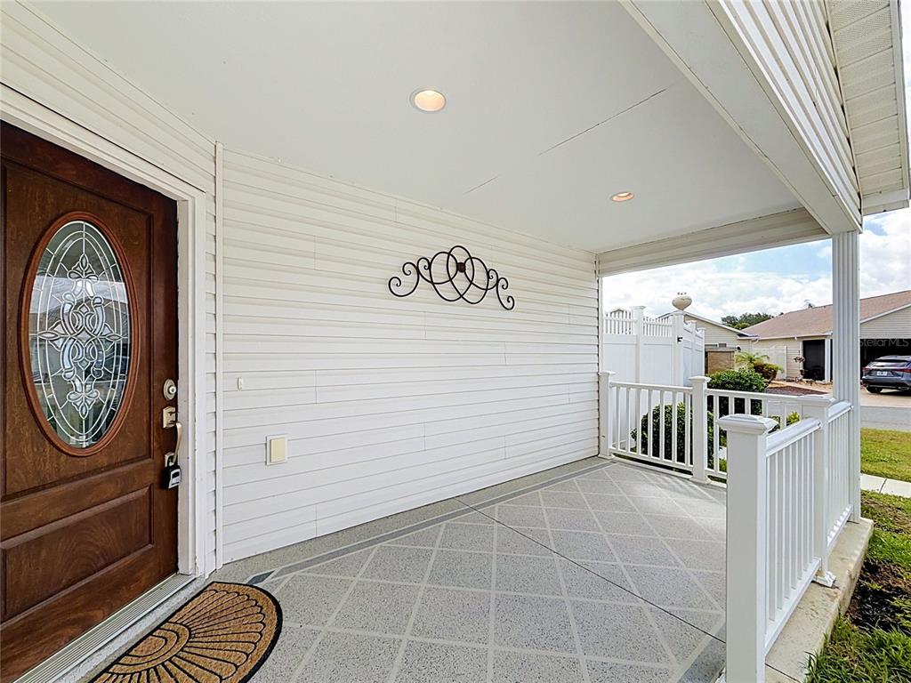 A covered front porch features white siding, a wooden front door, and a concrete floor — 2565 LINDEWOOD STREET, THE VILLAGES, FL