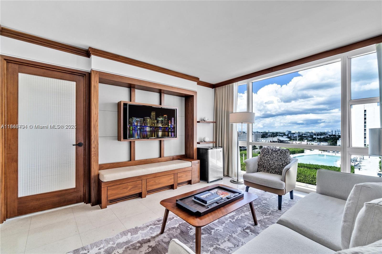 A modern living room features wood cabinetry, a wall-mounted television, and floor-to-ceiling windows overlooking a pool — 6801 Collins Ave # 818, Miami Beach FL 33141, MIAMI BEACH, FL
