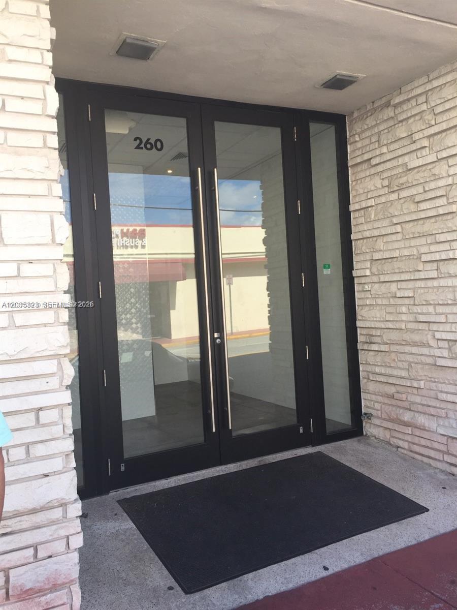 A glass double door entrance with a black frame is set between light-colored stone walls — SURFSIDE, FL