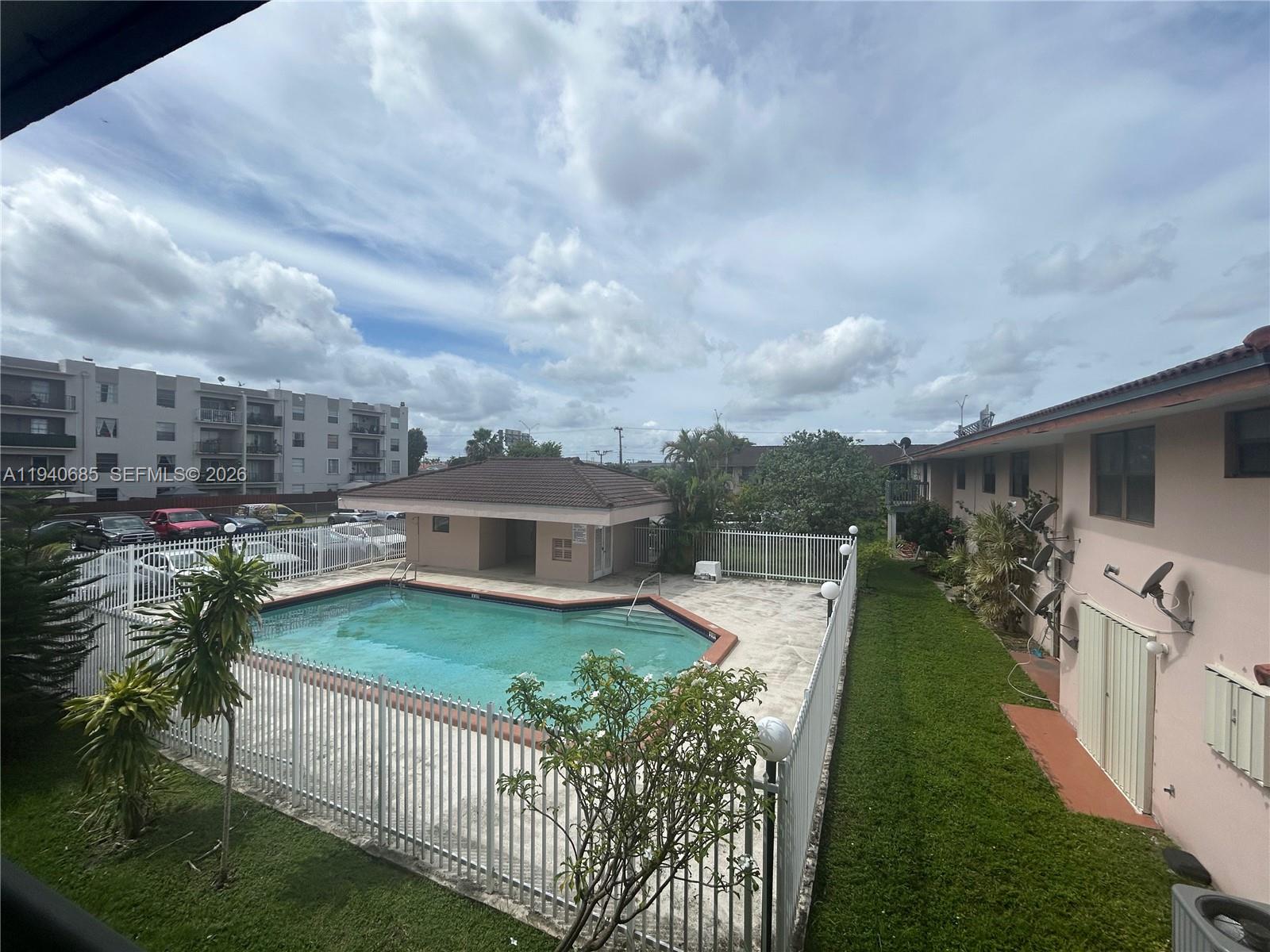 An outdoor swimming pool is enclosed by a white fence next to a residential building — 5851 W 20th Ave # 409, Hialeah FL 33012, HIALEAH, FL