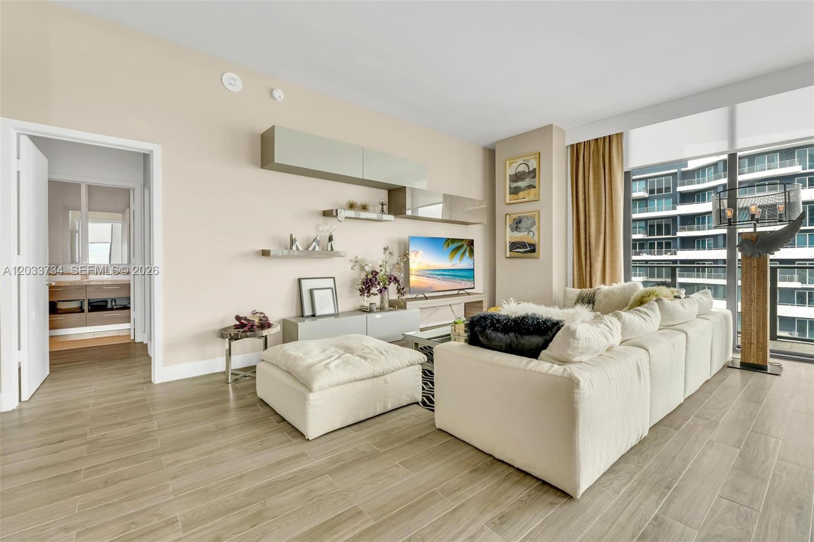A modern living room features a large white sectional, wall-mounted cabinetry, and floor-to-ceiling windows — 1300 S Miami Ave, Miami FL 33130, MIAMI, FL