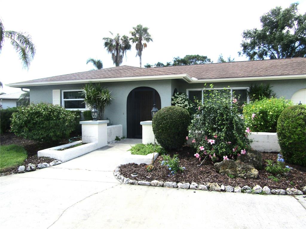 A single-story house features a gray exterior, a dark arched doorway, and landscaped front gardens — 4700 GREENCROFT ROAD, SARASOTA, FL