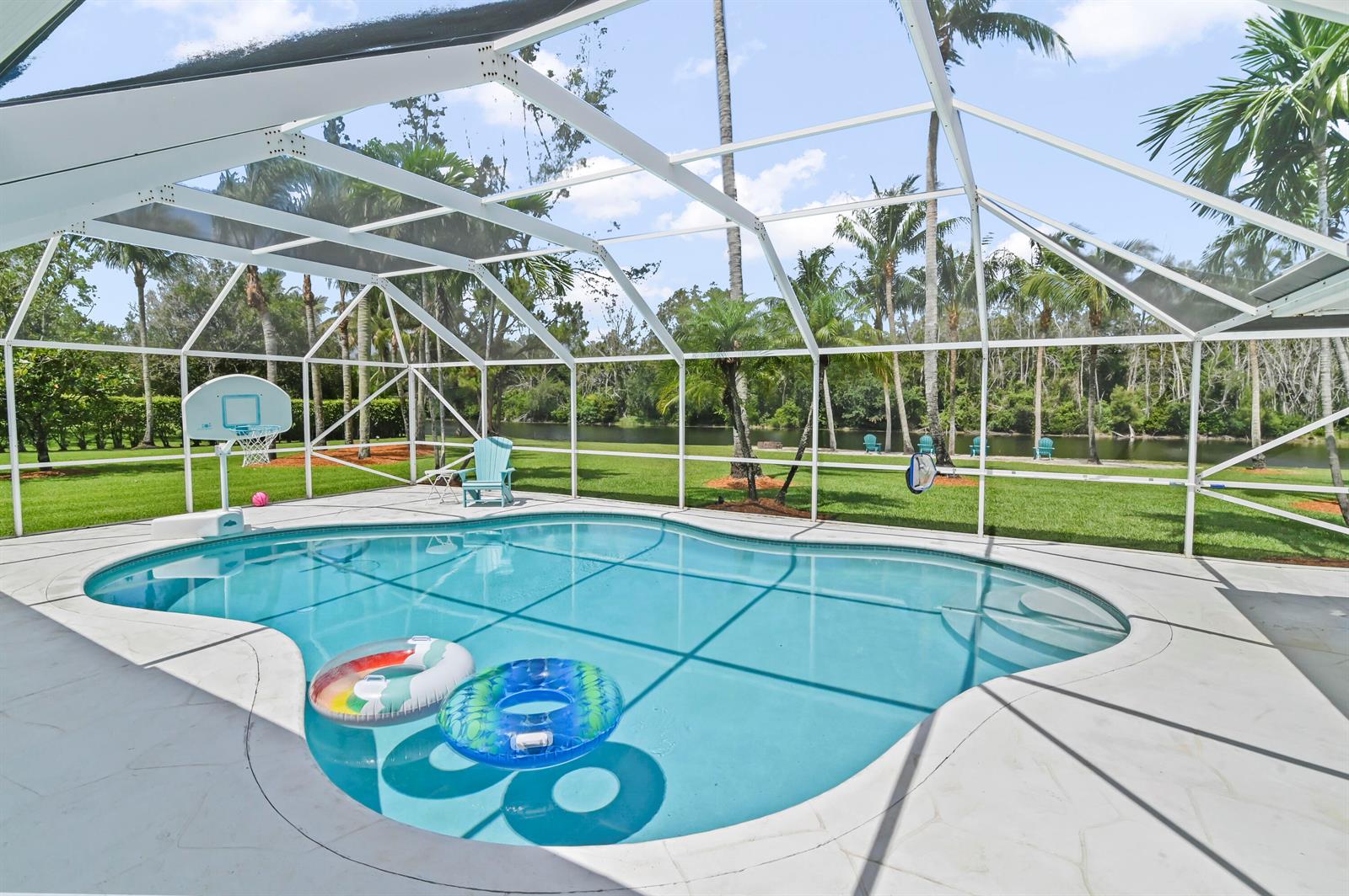 A swimming pool with two floats sits inside a screened lanai overlooking a grassy yard — 5637 Lago Del Sol Drive, Lake Worth FL 33449, LAKE WORTH, FL