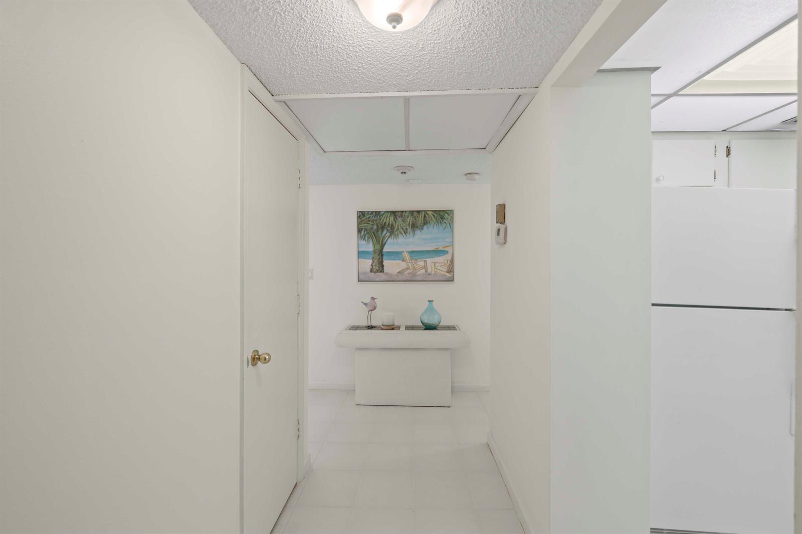 A white hallway leads to a console table with a beach-themed painting and decor — 951 De Soto Road # 232, Boca Raton FL 33432, BOCA RATON, FL