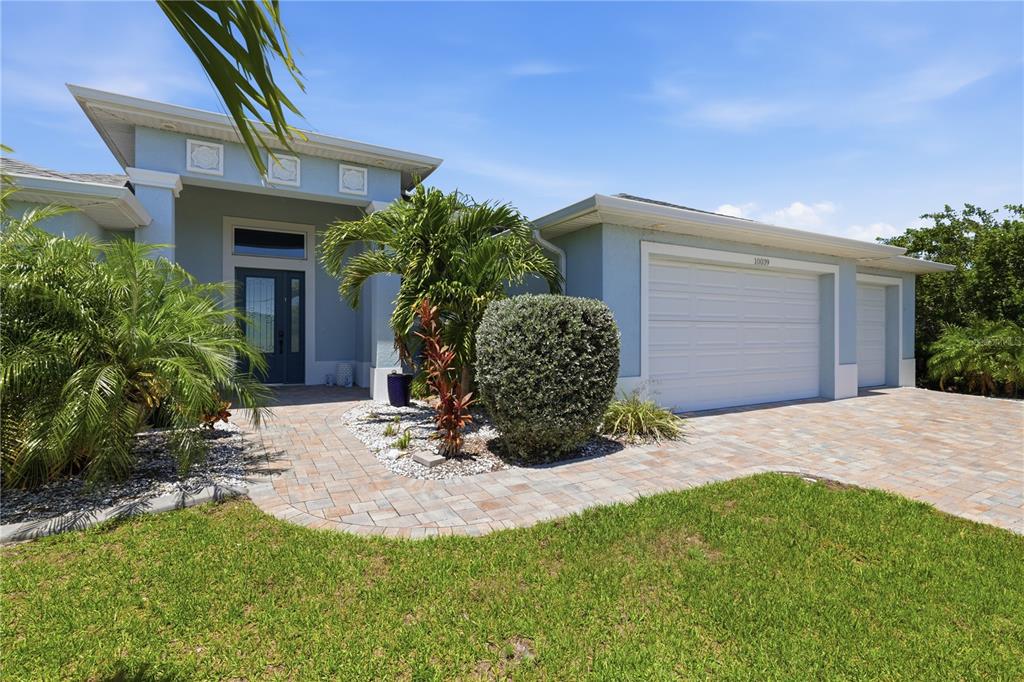 A light blue house features a paver walkway, a three-car garage, and tropical landscaping — 10039 KINGSVILLE DRIVE, PORT CHARLOTTE, FL