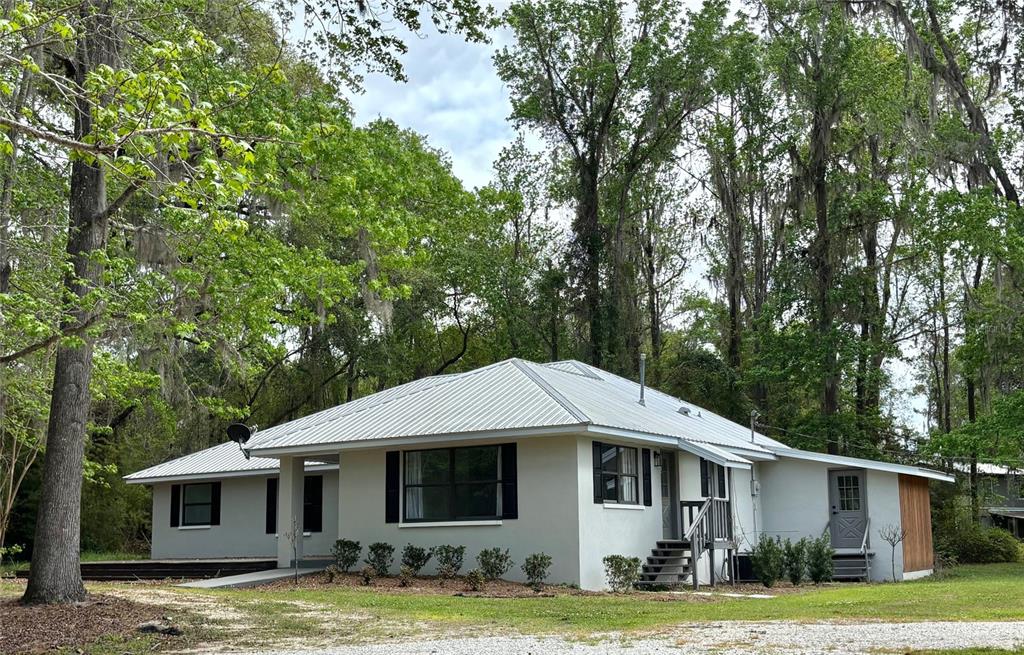 A single-story house with a metal roof and light-colored stucco exterior sits among tall trees — 18917 NW COUNTY ROAD 239, ALACHUA, FL