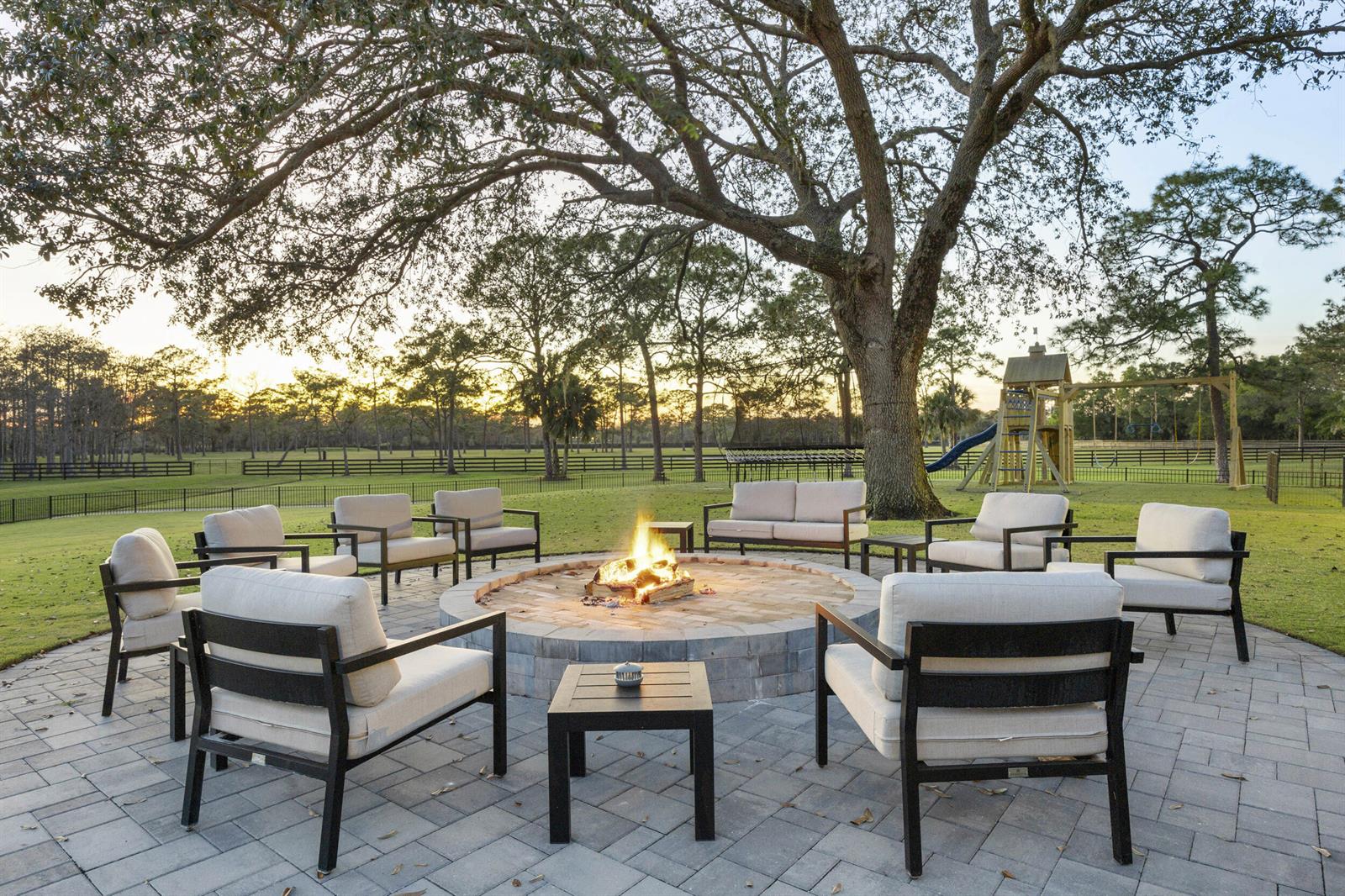 A stone fire pit surrounded by outdoor seating on a paved patio beneath a large tree — 20000 SE Mack Dairy Road SE, Jupiter FL 33478, JUPITER, FL