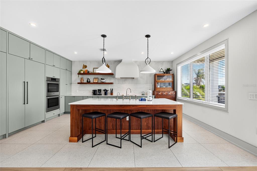 A modern kitchen features a wooden island with four stools, light green cabinetry, and white countertops — 100 12TH STREET N, BRADENTON BEACH, FL