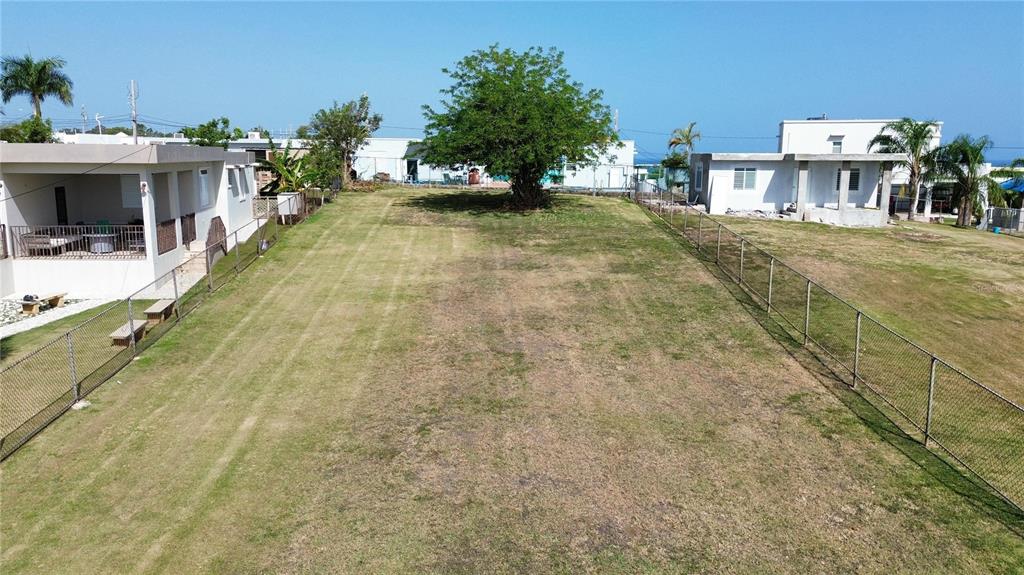 A large, grassy vacant lot is enclosed by chain-link fencing between two residential buildings — SOLAR 4 BO YEGUADA, CAMUY, PR