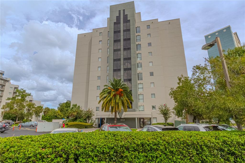 A tall, multi-story apartment building stands behind a manicured hedge and a parking lot — 6165 CARRIER DRIVE UNIT 2804, ORLANDO, FL